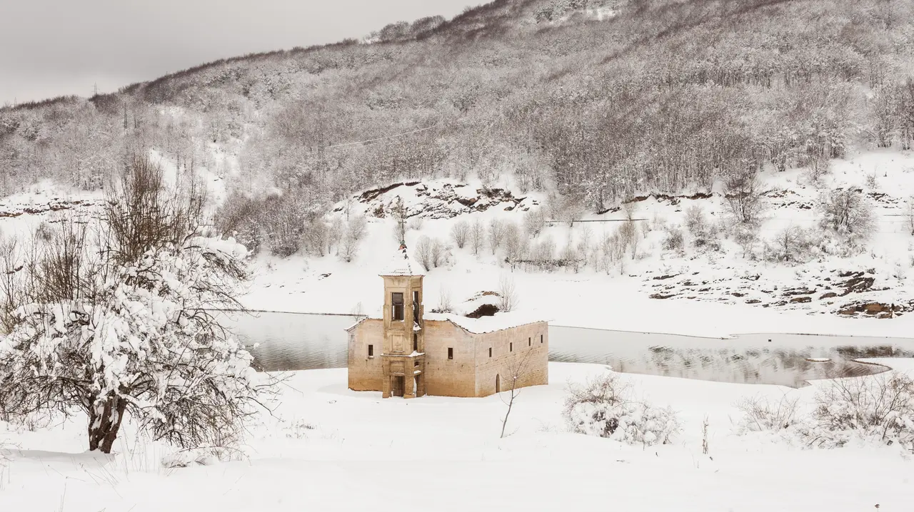 The half-submerged stone church of St. Nicholas standing in the snowy shoreline of Lake Mavrovo