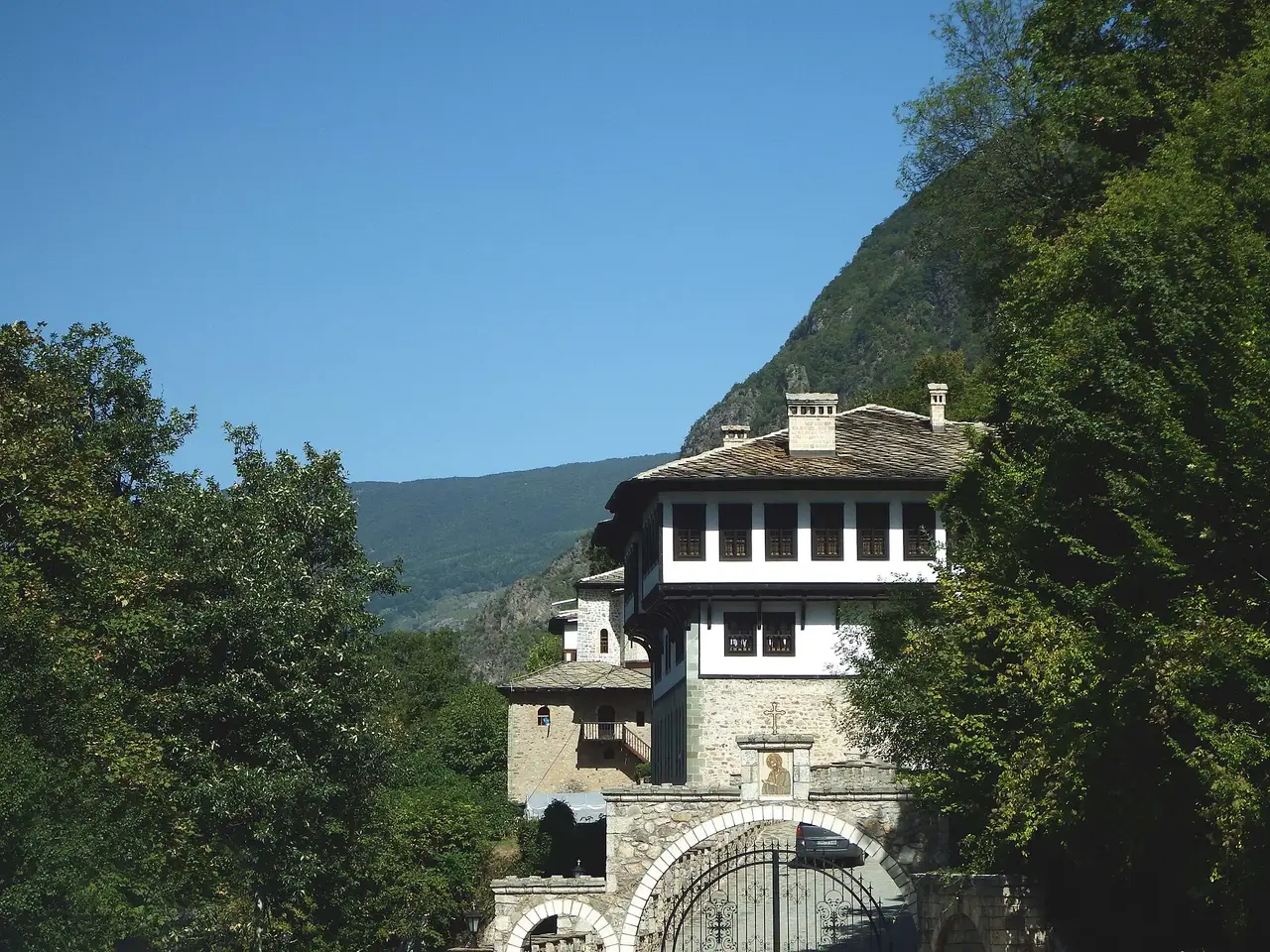 The white upper storeys and stone base of the Bigorski monastery lodging above an arched stone gate, with a forested mountainside behind