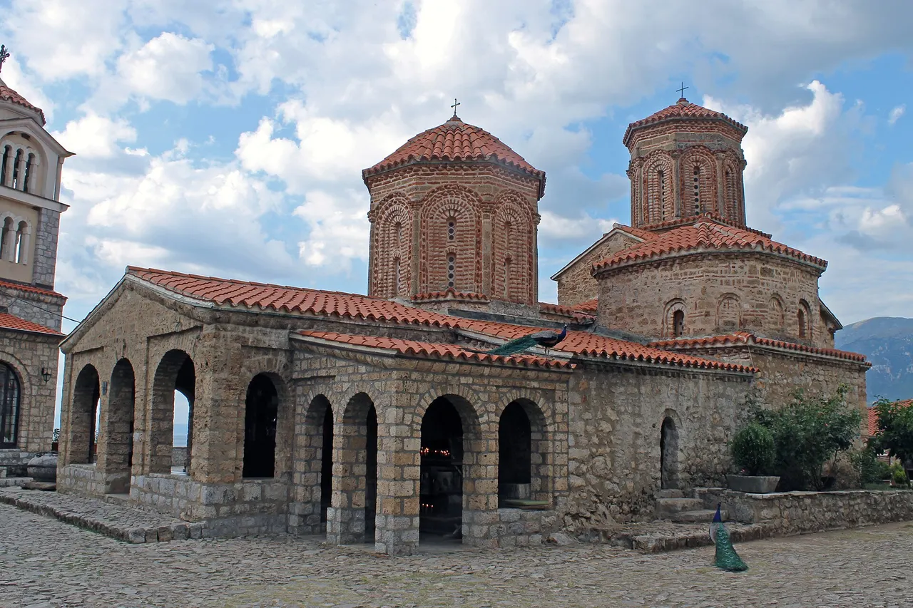 The stone church of Sveti Naum with two brick domes, an arcaded porch and red tiled roofs, a peacock perched on the roof and another on the cobbles