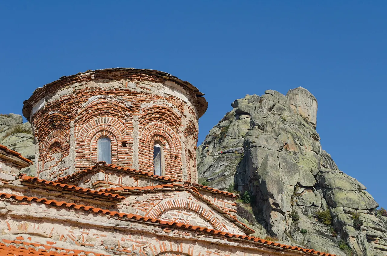 The brick and stone dome of the Treskavec church with red tiled roofs below and a tall rock outcrop behind it against a deep blue sky