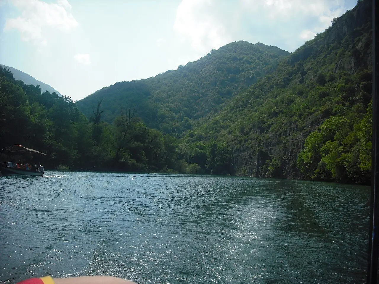 A tour boat with passengers on the green water of Matka Canyon between steep wooded cliffs near Skopje