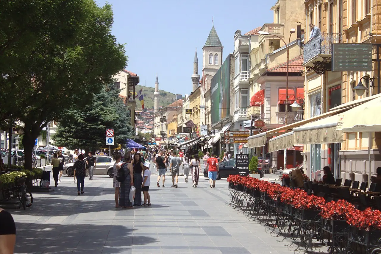 People strolling along Sirok Sokak, Bitola's pedestrian street, past cafe terraces with red flower boxes and 19th-century facades on a sunny day