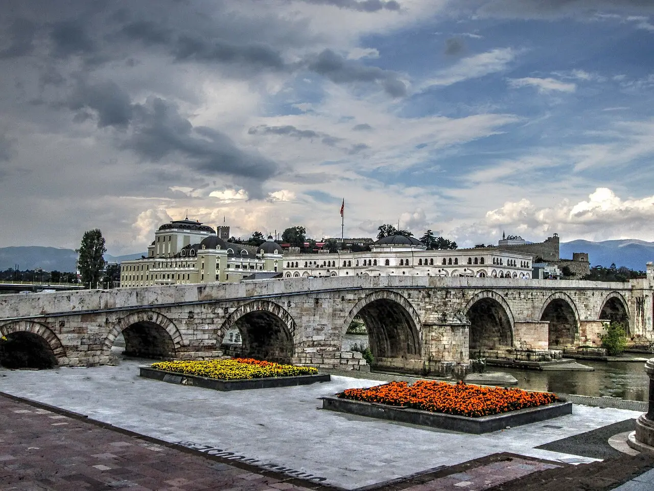 Skopje's Ottoman-era Stone Bridge over the Vardar river, with flower beds, government buildings and the Kale fortress behind under a dramatic sky