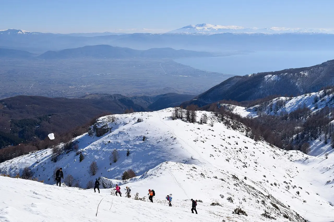 A line of hikers climbing a snow-covered ridge in North Macedonia with a wide valley far below
