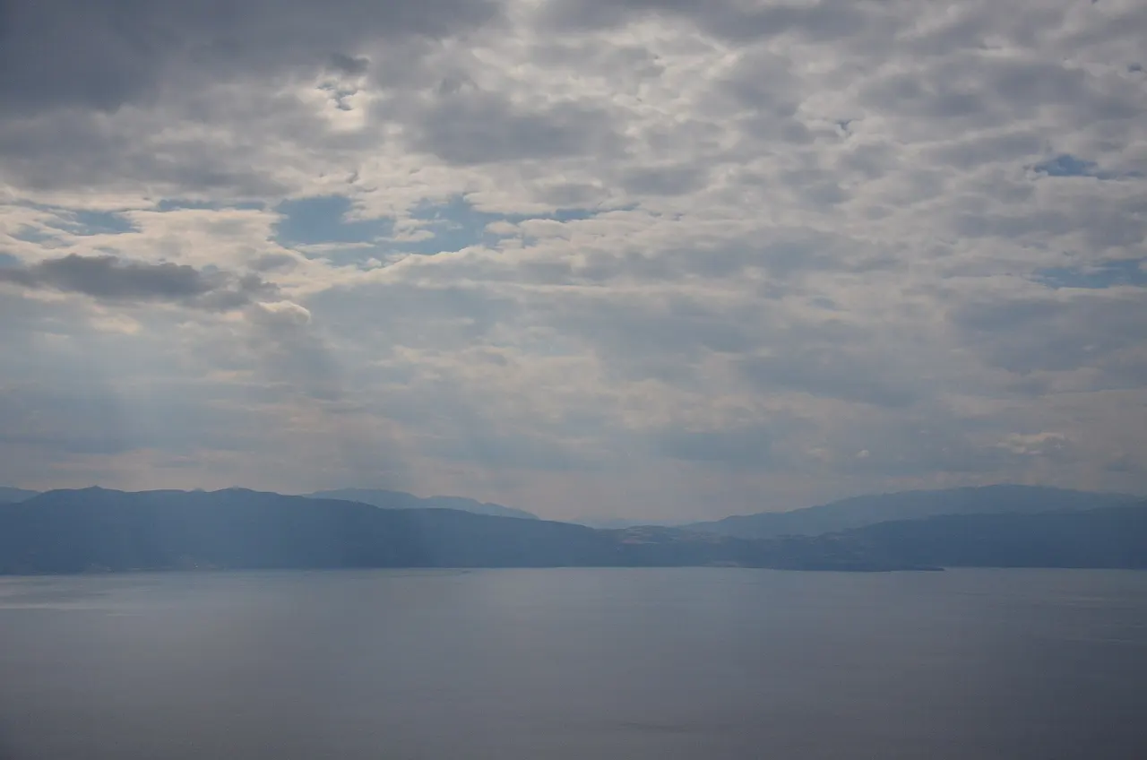 Lake Ohrid under a heavy cloud sheet, with mountains along the far shore and light breaking on the water