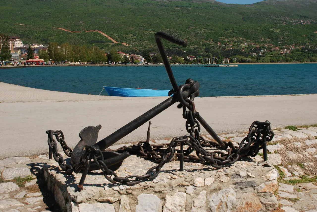 A black iron anchor on the Ohrid waterfront promenade with turquoise lake water and red-roofed houses on the far shore