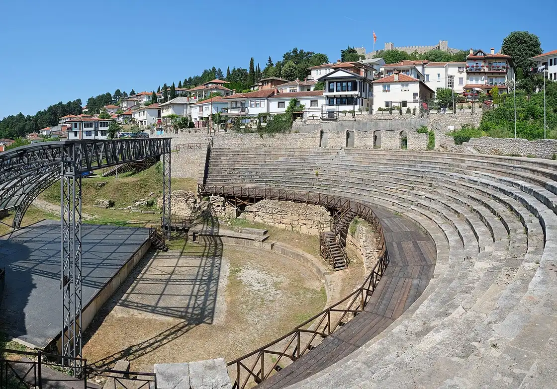 The curved stone tiers of the ancient theatre in Ohrid, a modern stage platform below and the old-town houses and a hilltop fortress rising behind