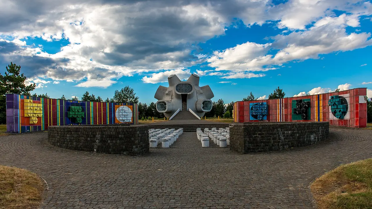 The white Makedonium monument with its bulbous window pods, flanked by long walls of coloured mosaic panels under a cloudy sky