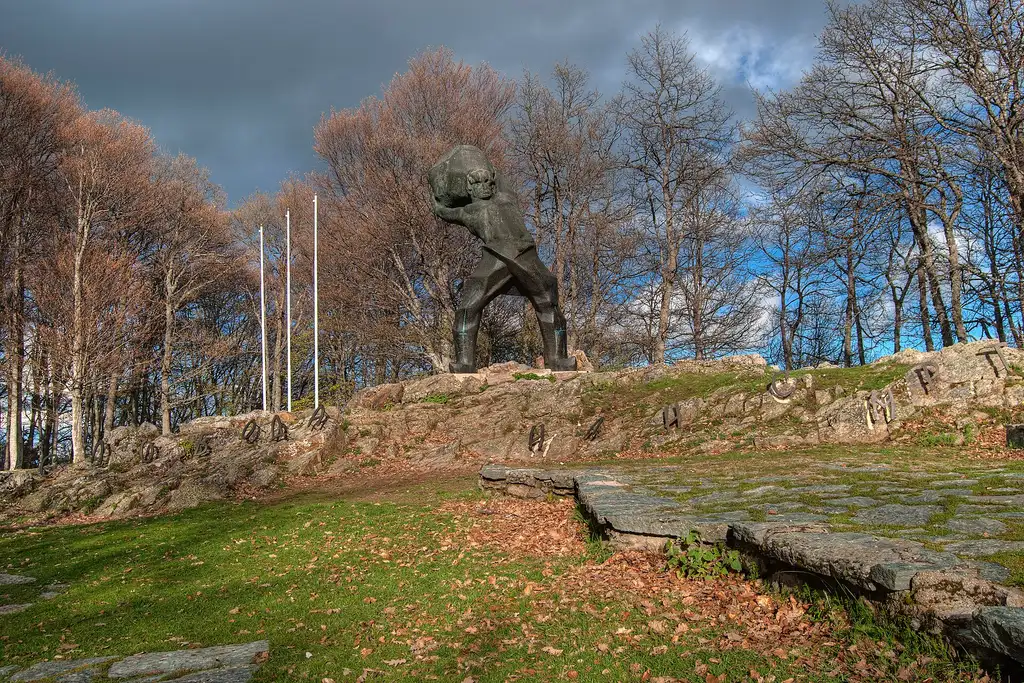 A dark bronze statue of a fighter lifting a boulder on a rocky outcrop among bare autumn trees at Mečkin Kamen above Kruševo