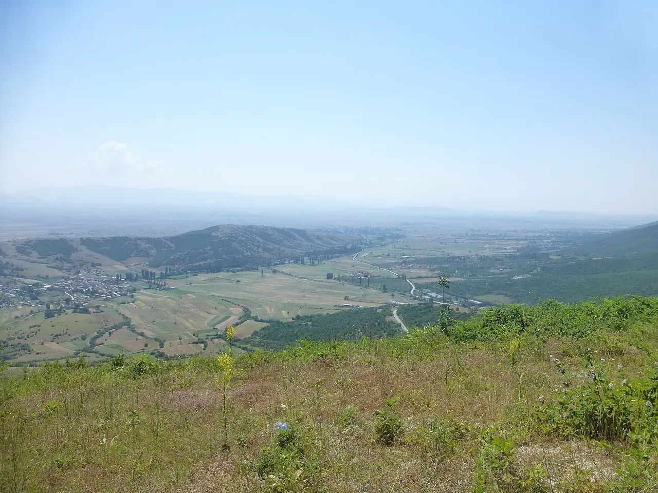 A grassy roadside slope looking south-east over a patchwork of fields, a village on the left and the hazy Pelagonia plain beyond
