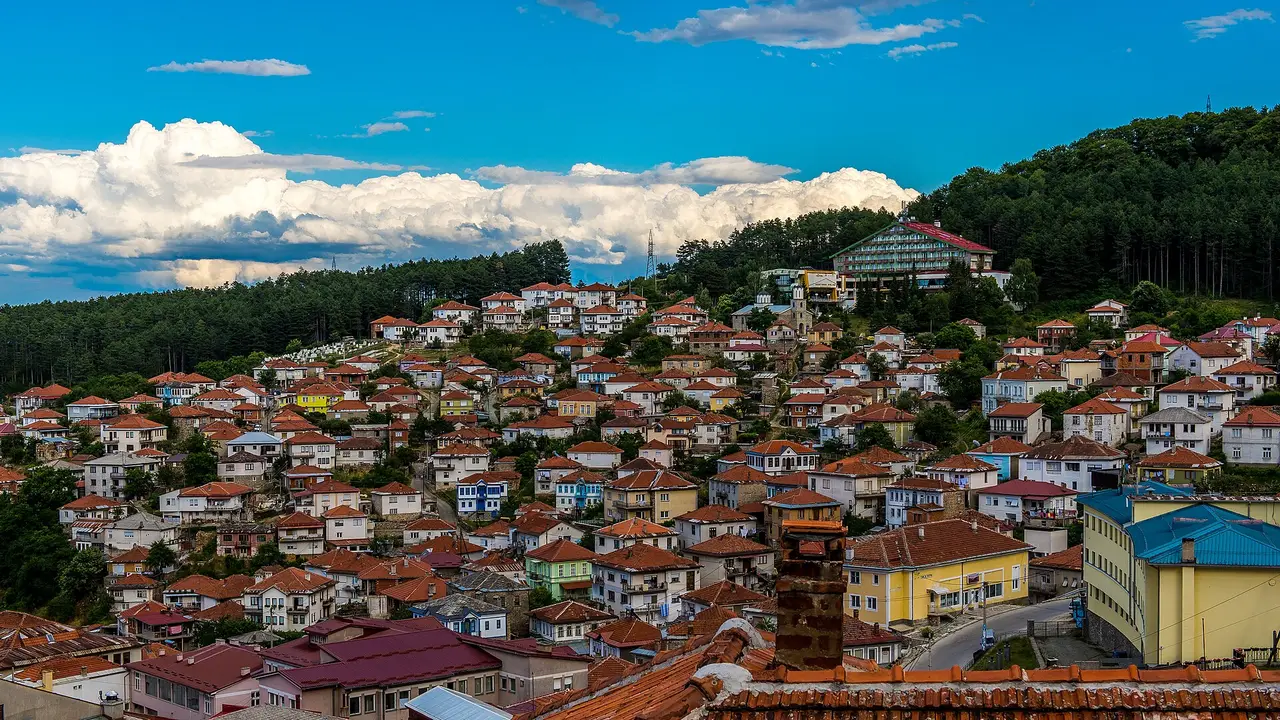 Red-roofed houses of Kruševo stacked up a steep hillside under pine forest and summer clouds