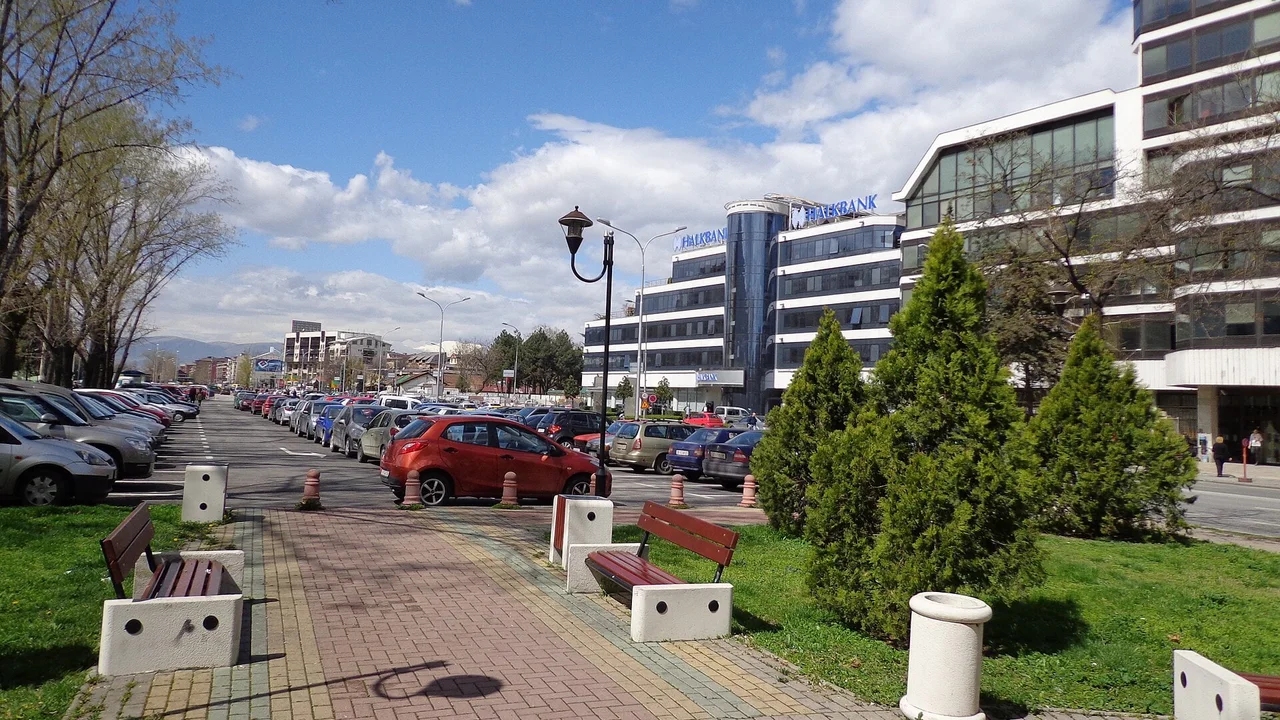 Rows of cars parked along a street beside a paved park path with benches in central Skopje