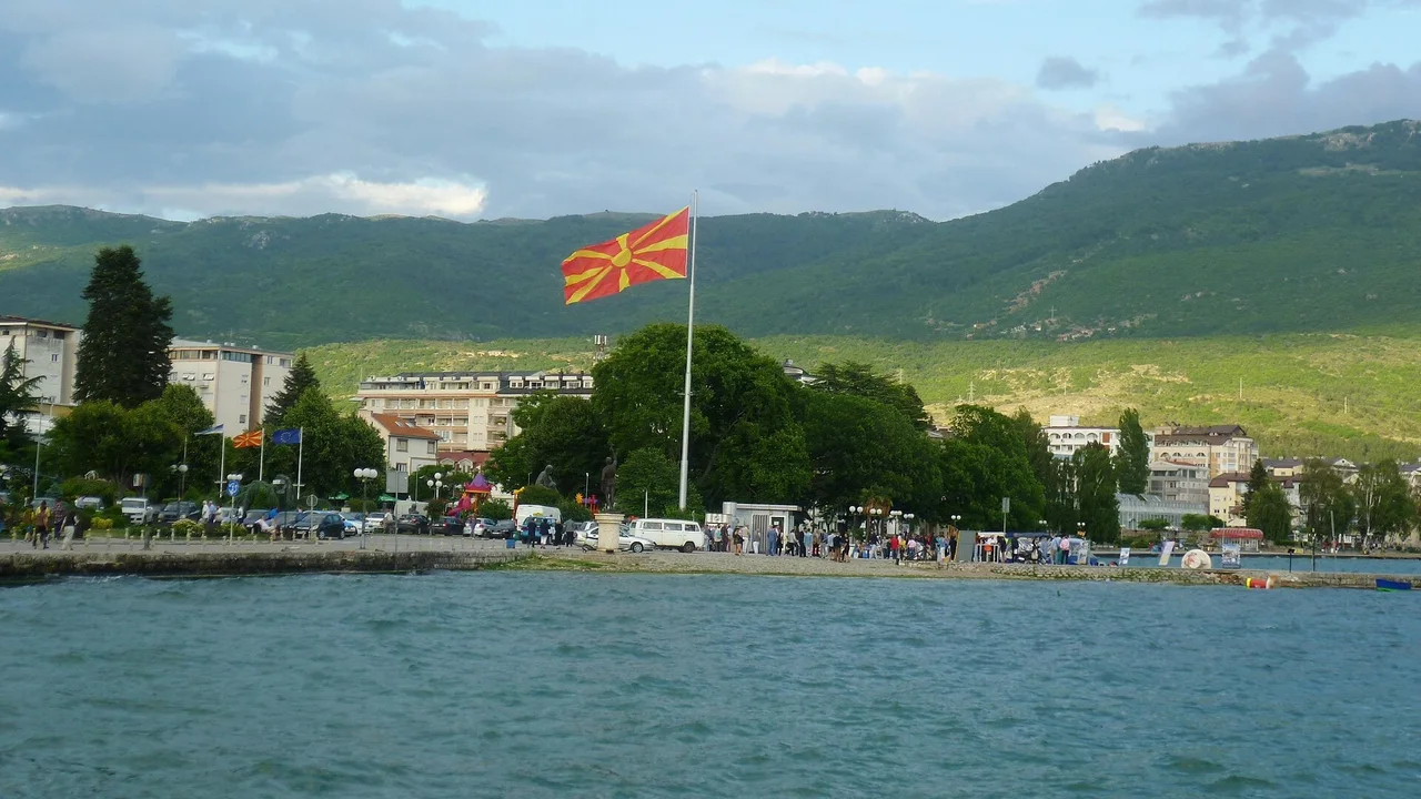 Cars parked along the quay beside the tall flagpole on the harbour front in Ohrid, seen from the lake with green hills behind