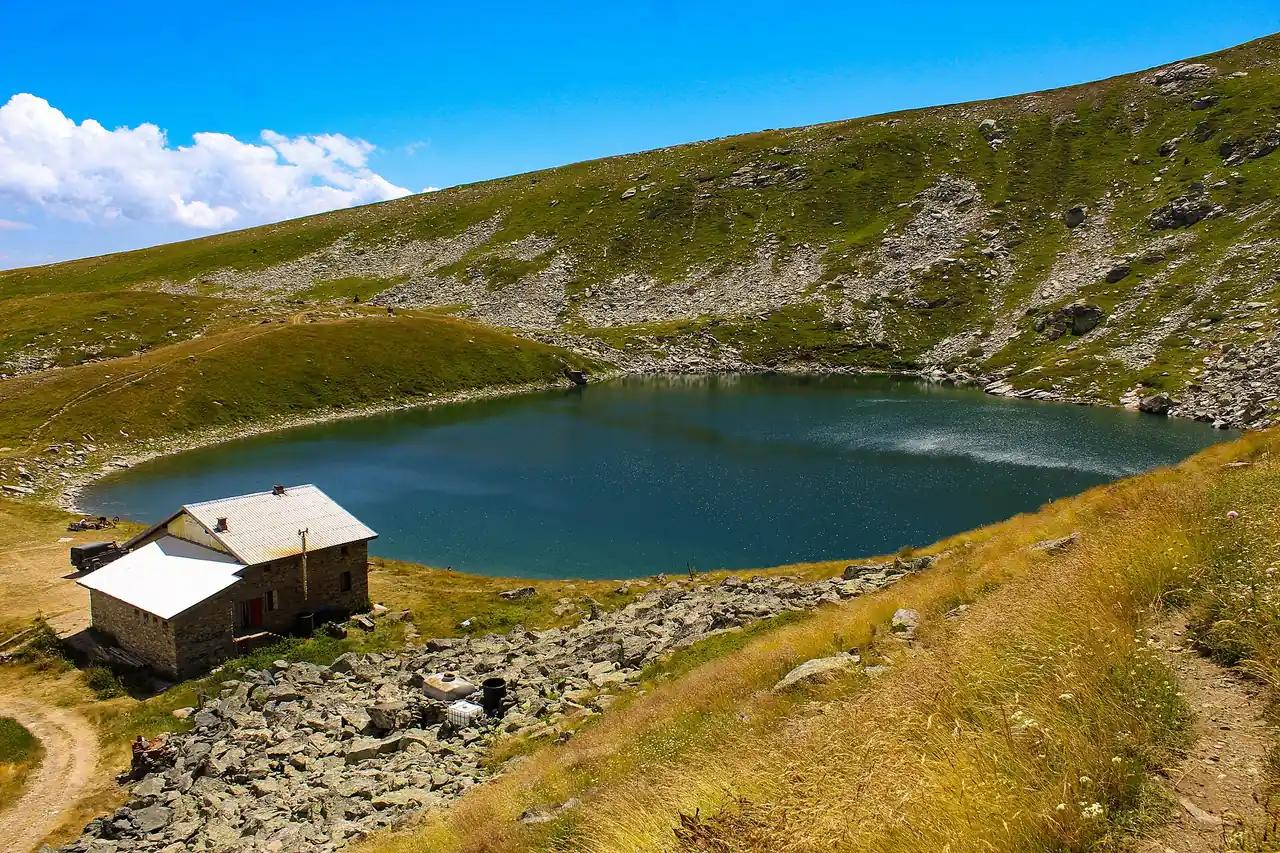 The deep blue Golemo Ezero glacial lake below rocky Baba slopes, with a stone mountain hut on its shore