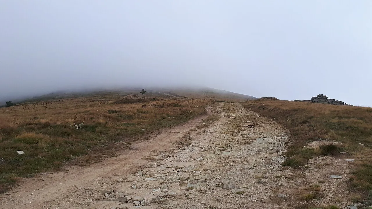 A stony mountain path climbing toward a grassy ridge that disappears into low cloud on Pelister