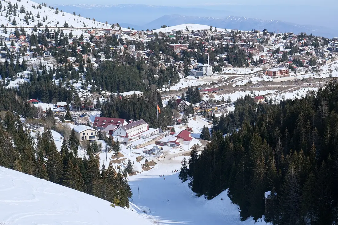 Popova Šapka's chalets, hotels and a minaret spread across a snowy hillside among pine trees, seen from above the ski run