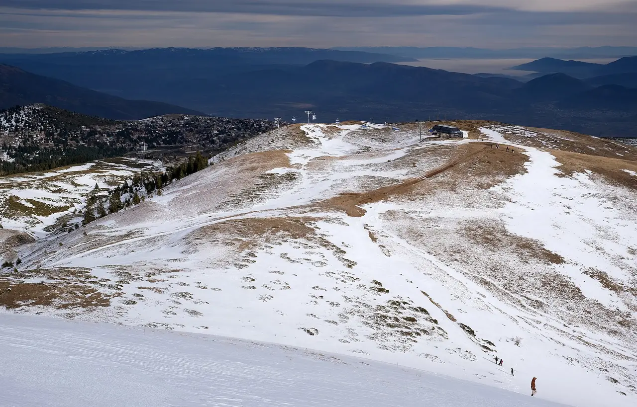 Patchy snow and brown grass on the upper slopes of Popova Šapka near a lift top station, with the Polog valley in haze below