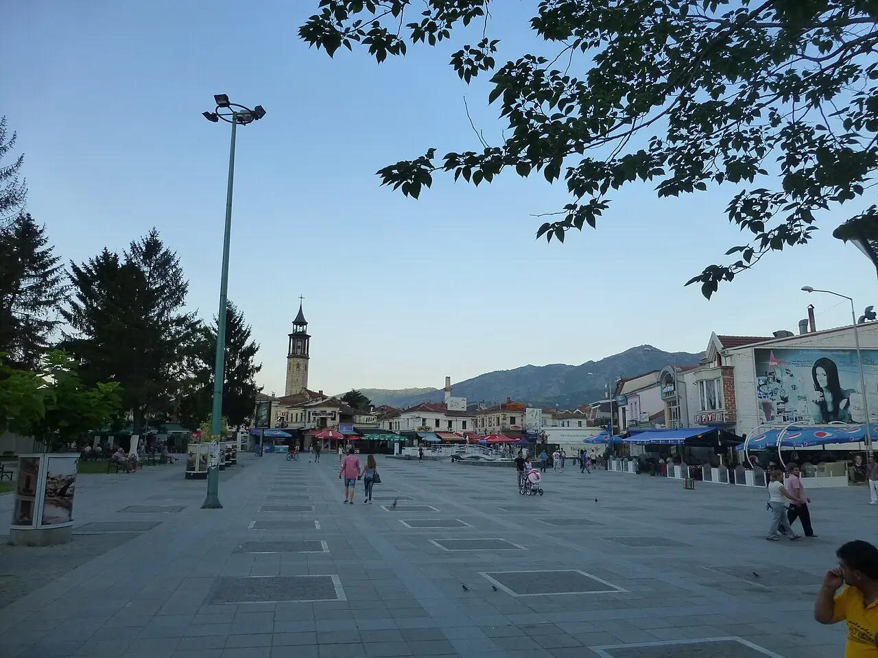 Prilep's broad pedestrian central square at dusk, with the stone clock tower at the far end and mountains behind