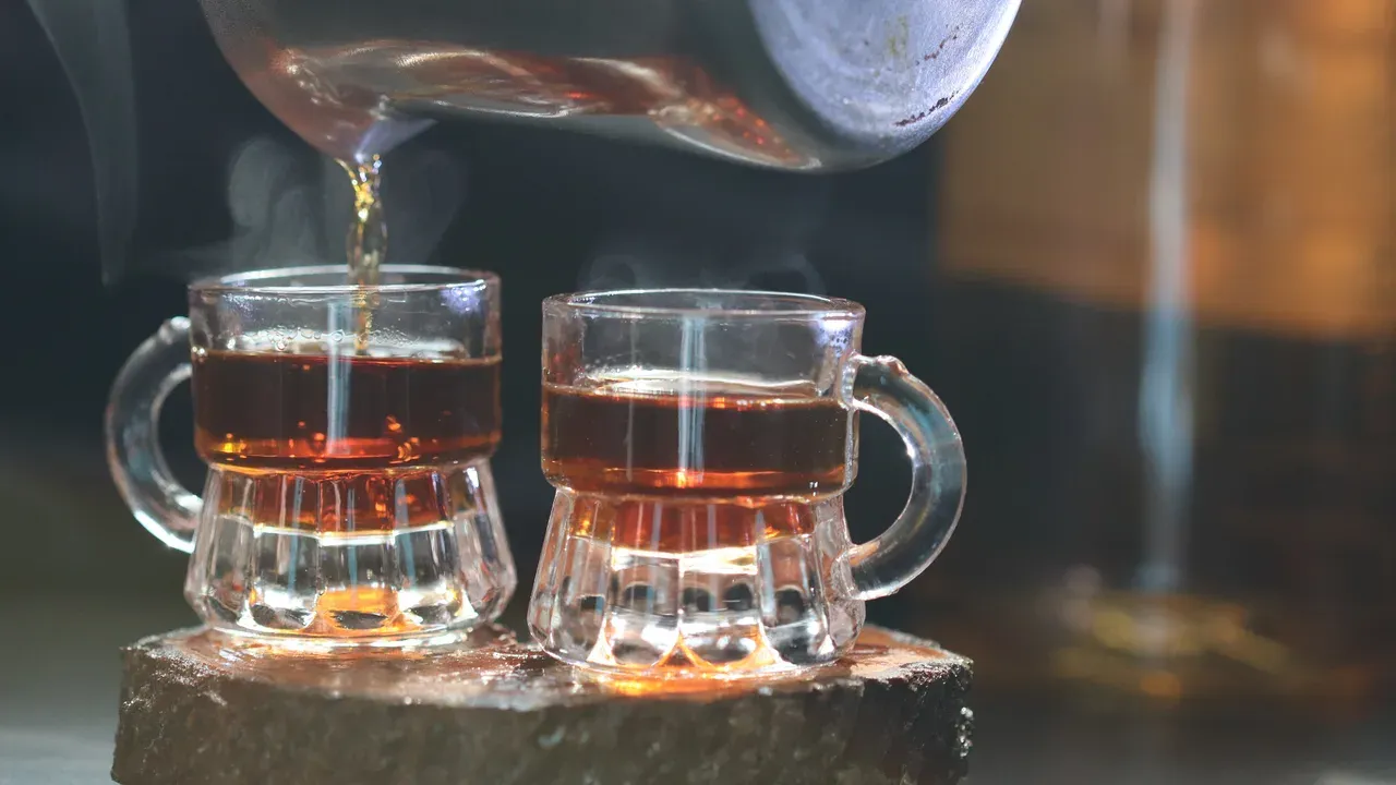Two small glass mugs of warm amber rakija being filled from a metal pot, steam rising from the glasses