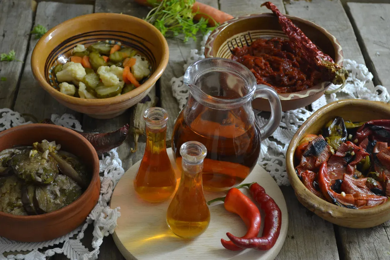 A glass jug and two small carafes of amber rakija on a wooden table among bowls of pickled vegetables, dried peppers and roasted aubergine