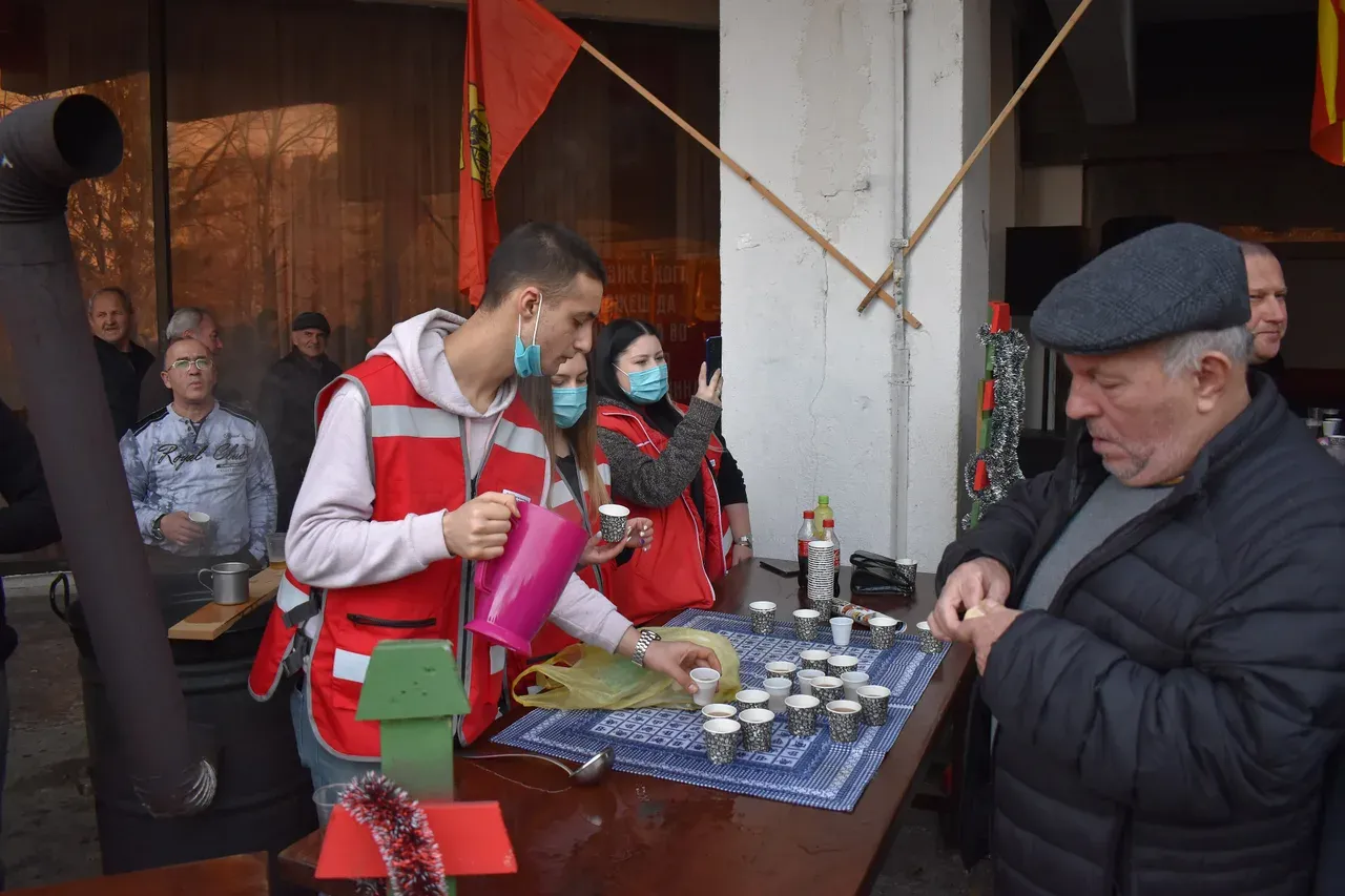 A volunteer in a red vest pouring from a pink jug at a table of small cups while an older man in a flat cap looks on