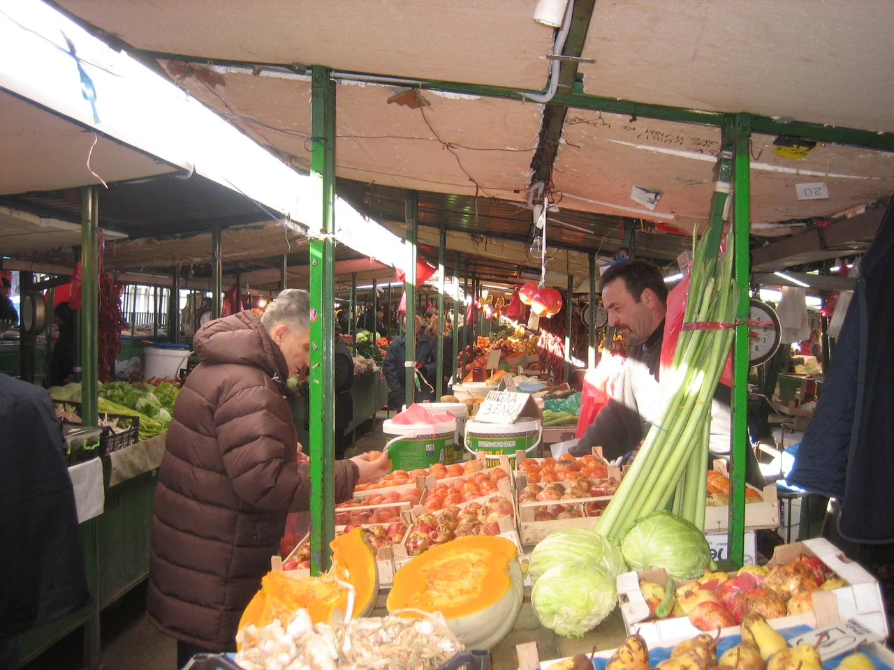 A shopper in a winter coat picks over fruit at a produce stall in Skopje's Bit Pazar, with cabbages, pumpkin and spring onions in the foreground