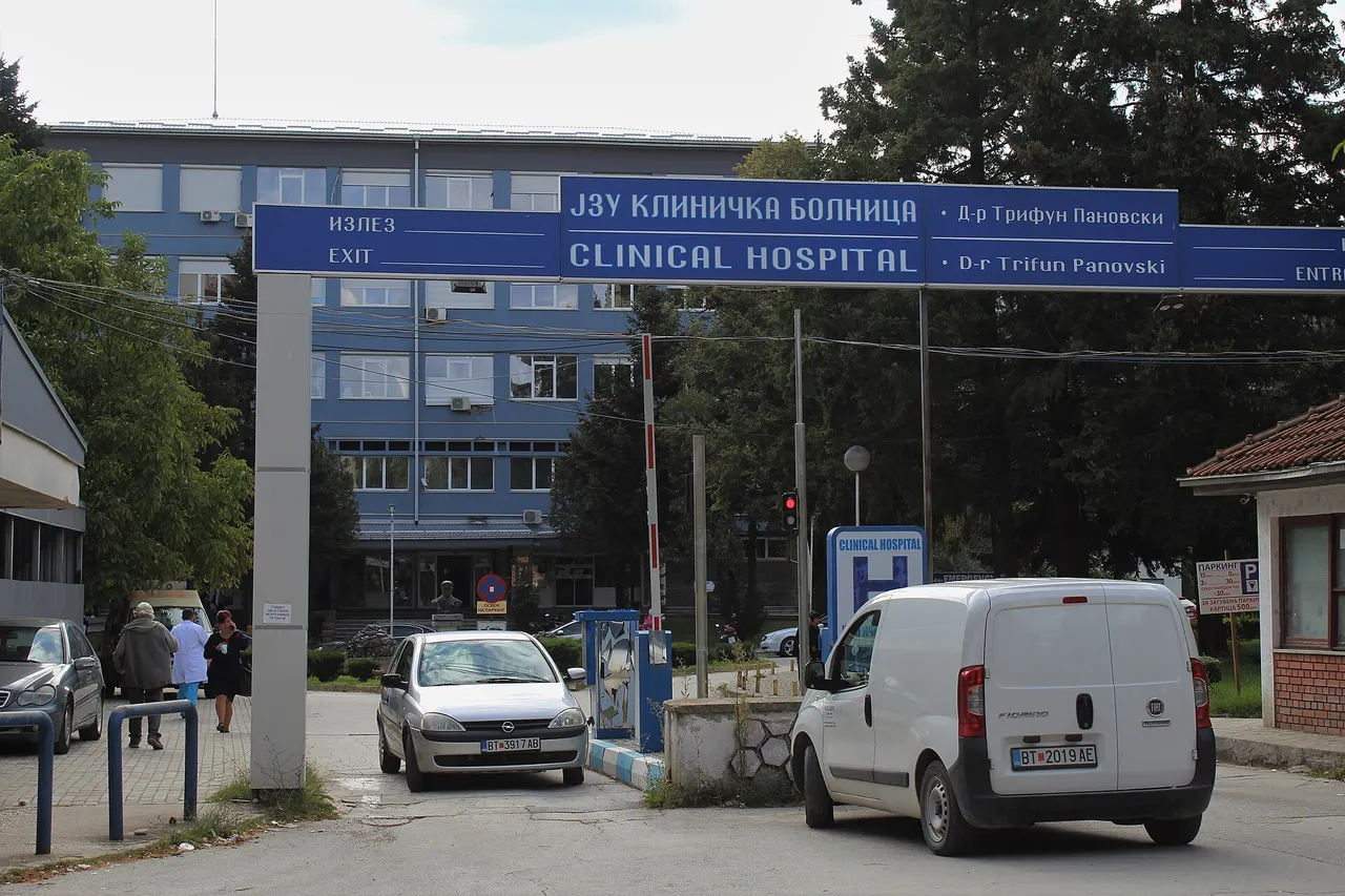 The entrance of the Clinical Hospital Dr. Trifun Panovski in Bitola, with a blue ward block behind a sign gantry, a barrier, parked cars and two people in white coats at the left