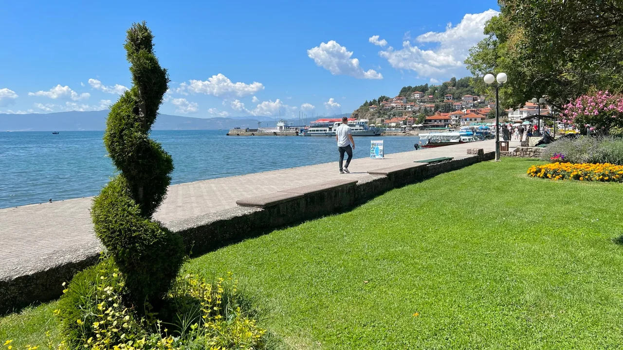 A man walks along the Lake Ohrid promenade on a sunny day, with a clipped evergreen and a wide lawn in the foreground and the old town on its hill across the water