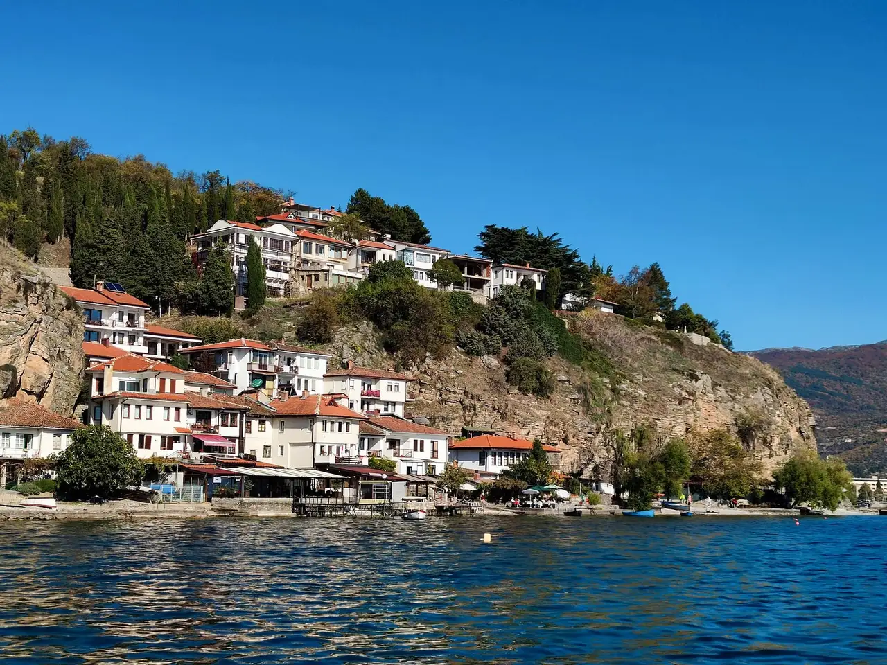 White houses with red-tiled roofs climb a rocky hillside above the blue water of Lake Ohrid, with lakeside restaurants at the water's edge