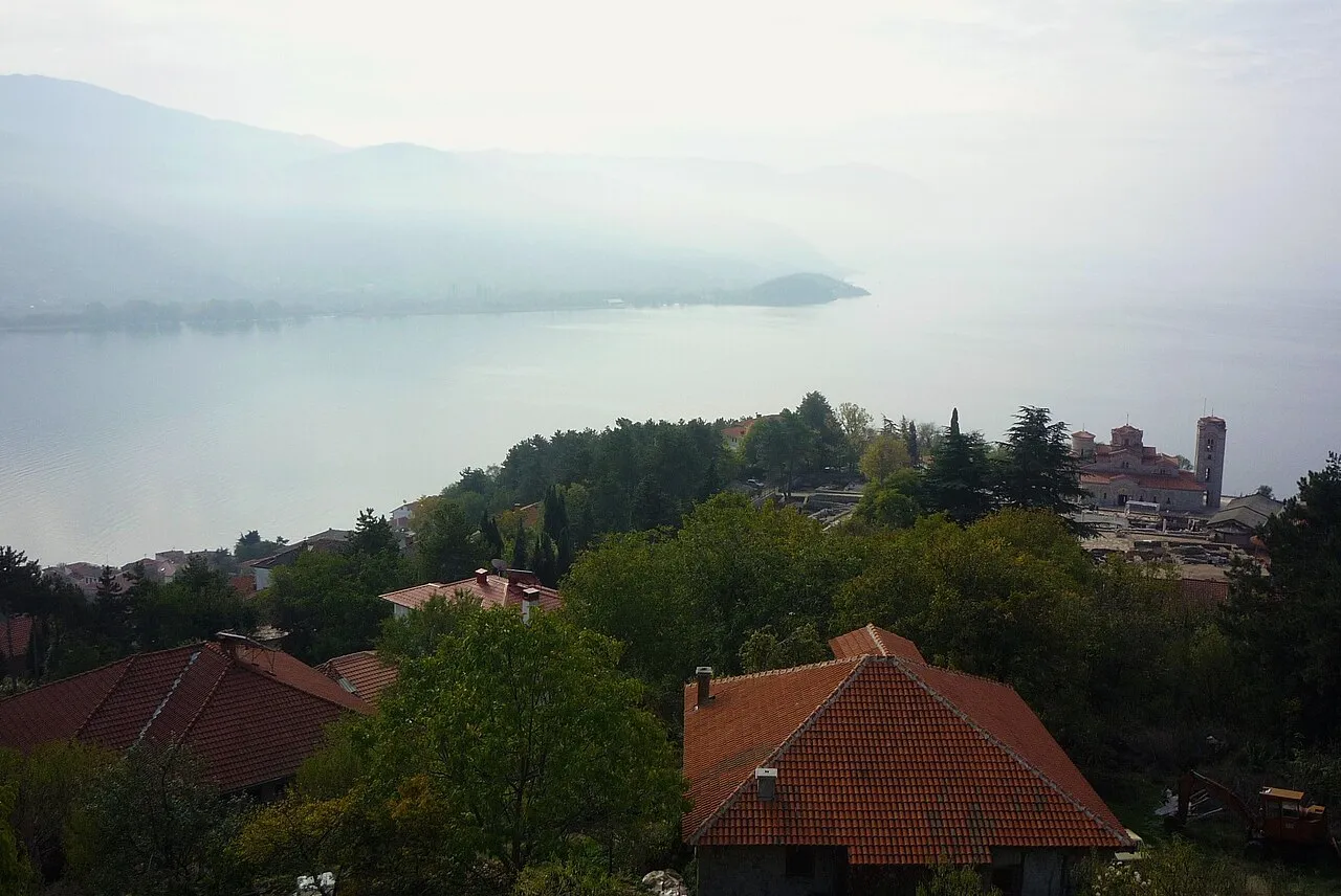 Hazy view from the fortress over red rooftops and trees to Lake Ohrid, with the Plaošnik church complex on a wooded point