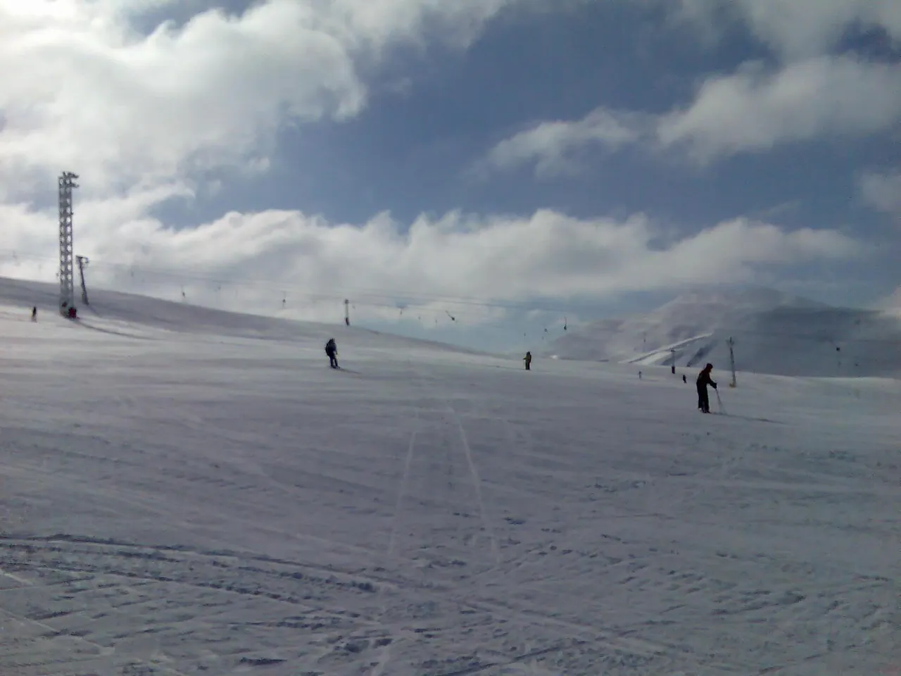 A few skiers on a wide open snow slope at Mavrovo with a lift pylon and a line of drag lifts under a cloudy sky