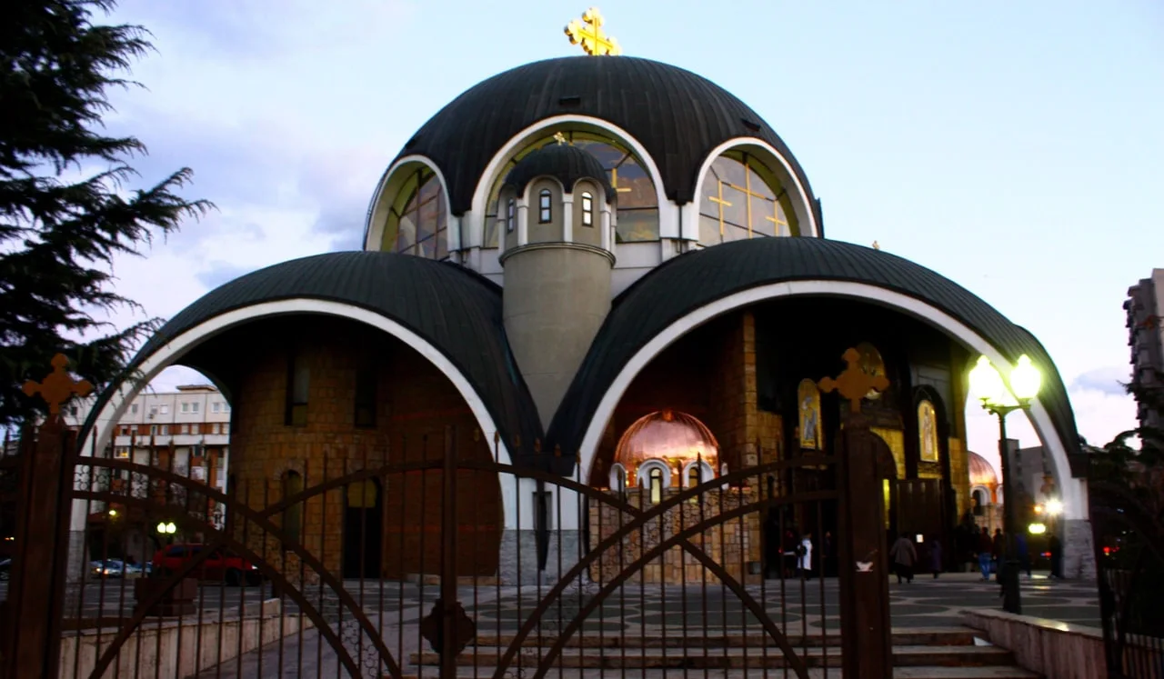 The domed Cathedral of St Clement of Ohrid in Skopje at dusk, seen through an iron gate, with lamps lit by the entrance