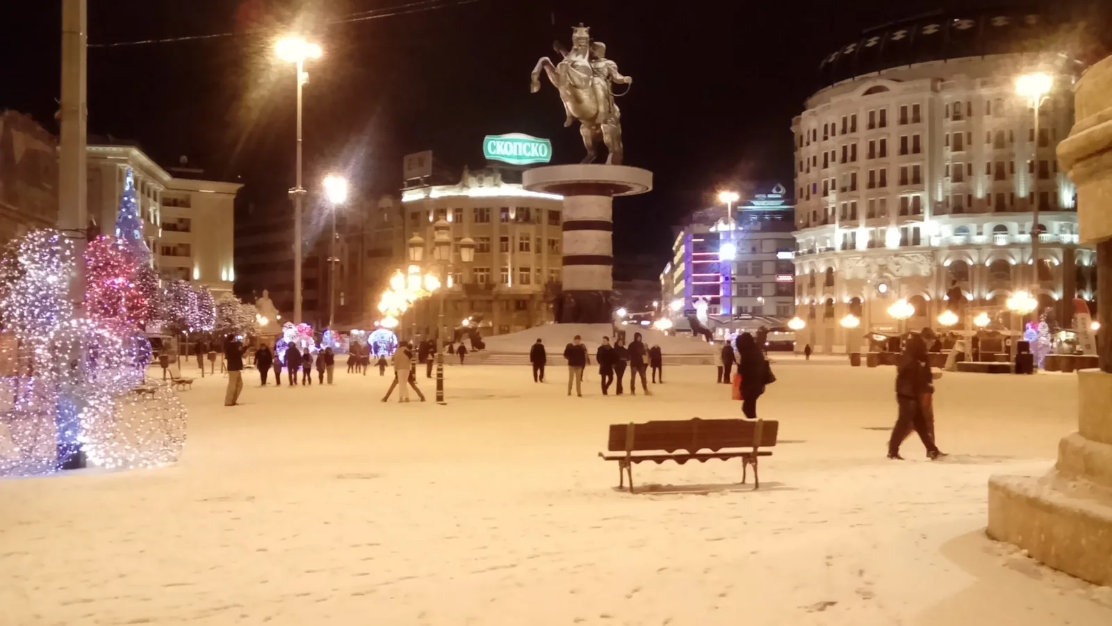 Macedonia Square in Skopje at night under fresh snow, with string lights, strollers and the Warrior on a Horse monument