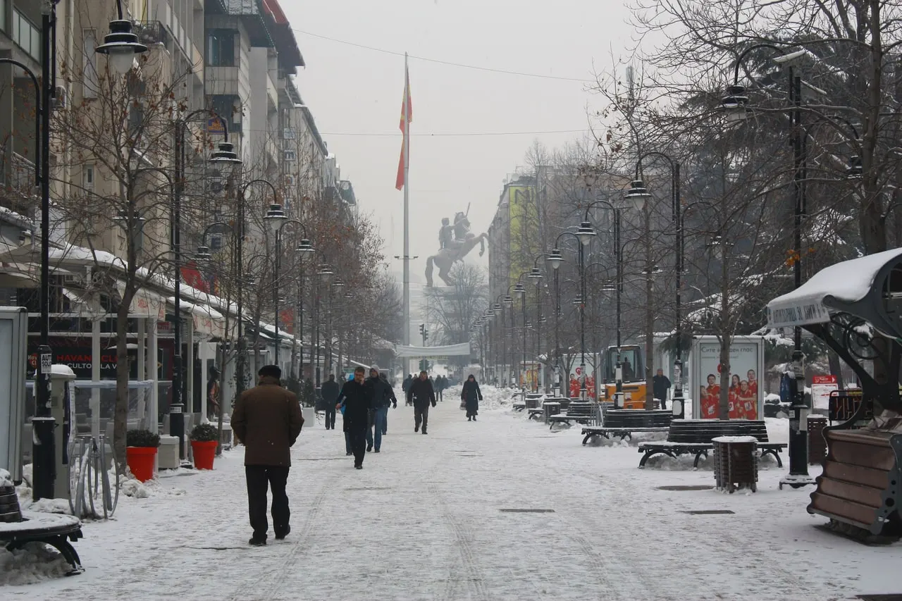 Snow-covered pedestrian Makedonija Street in Skopje with people walking, bare trees and the Warrior on a Horse monument at the far end