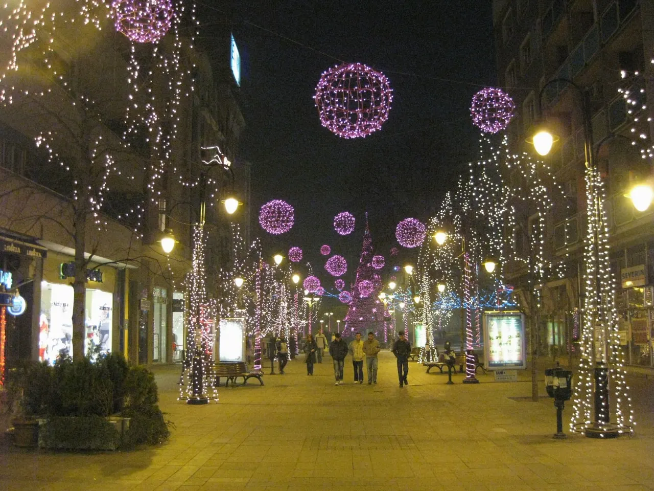 Makedonija Street in Skopje at night, lined with trees wrapped in white lights and hung with pink light globes, with a lit tree at the far end and a few people walking