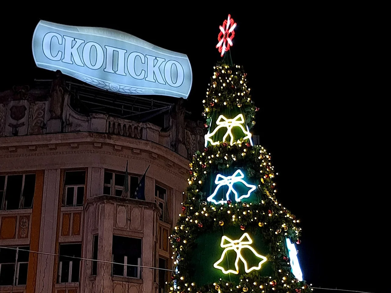 A tall Christmas tree on Macedonia Square covered in string lights and glowing bell shapes, with a lit Skopsko sign on the rooftop behind it