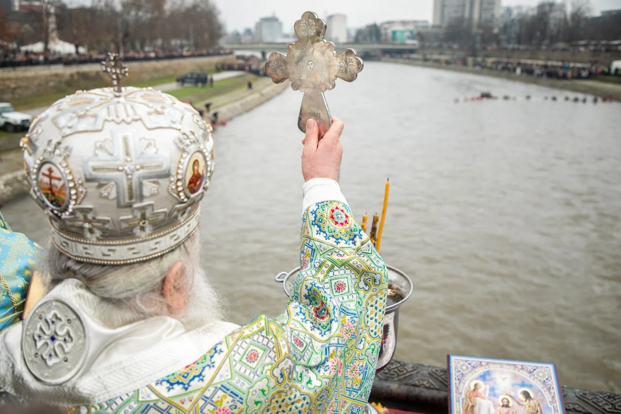 A priest in a white mitre and embroidered vestments holds a silver cross up over the Vardar river in Skopje, with crowds lining both banks