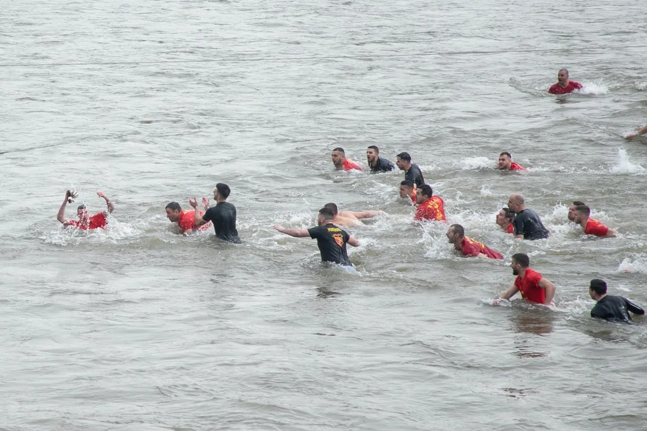 Dozens of men in red and black shirts swimming and wading in the grey Vardar river during the Vodici cross race