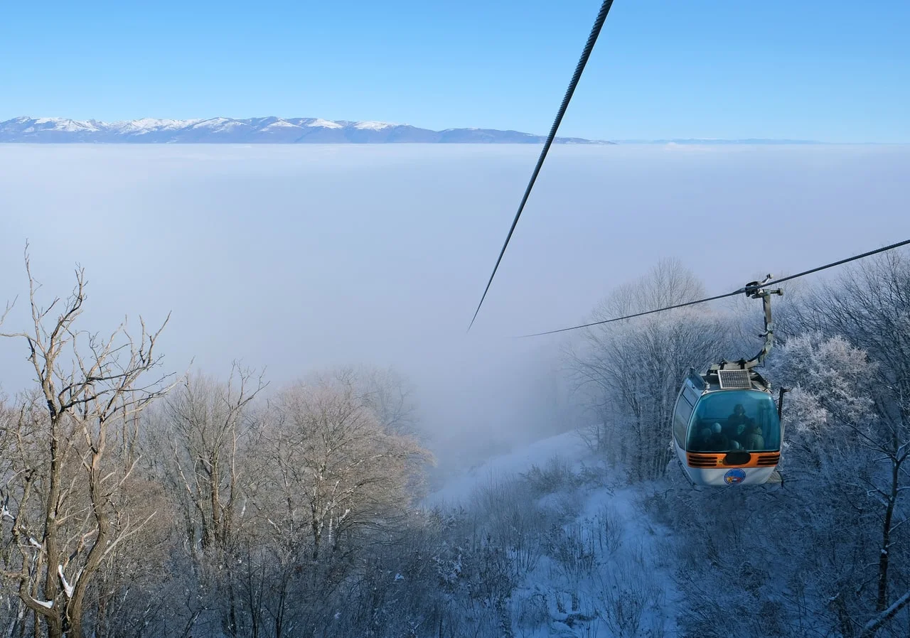 A cable car cabin above a sea of fog that fills the Skopje valley, with frosted trees below and snowy mountains on the horizon