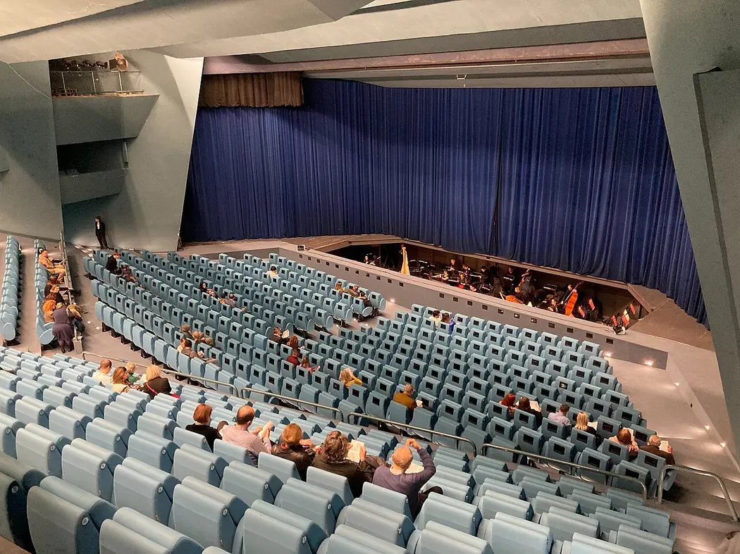A high-angle view inside the National Opera and Ballet auditorium in Skopje, tiered blue seats facing a stage with a blue curtain and an orchestra tuning up in the pit below