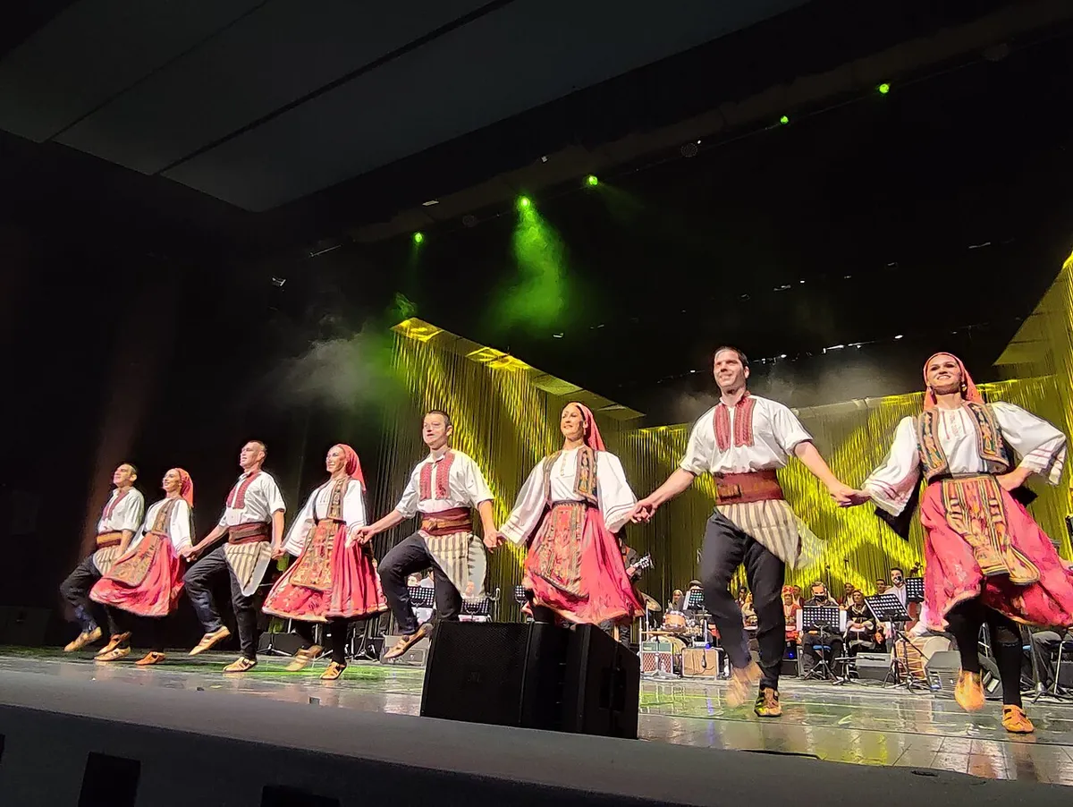 Dancers in traditional Macedonian costume performing on stage at the National Opera and Ballet, with musicians visible in the pit below