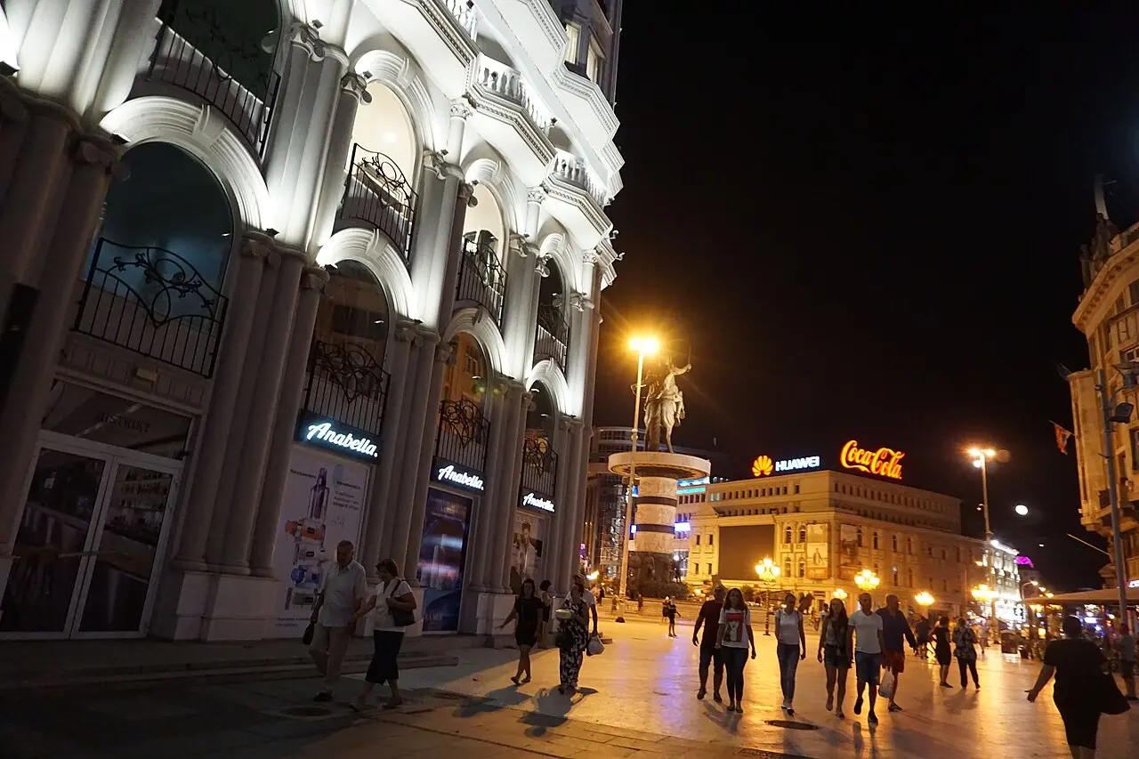 Pedestrians on a lit central Skopje street at night with neon signs