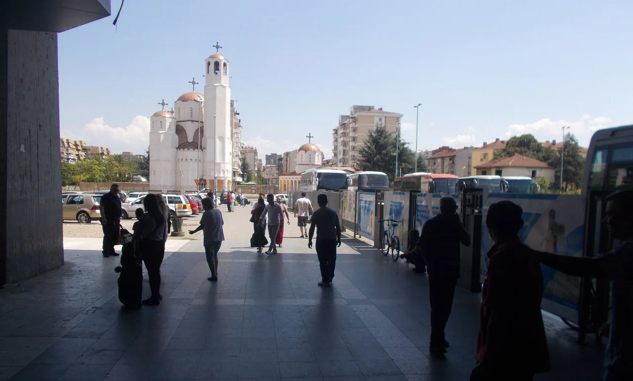 Passengers walking out of the shade of the Skopje bus terminus, with coaches lined up on the right and a white church in the distance