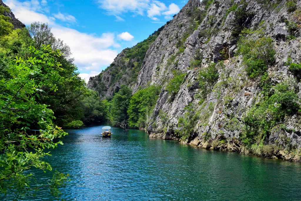 A small covered boat on the green water of Matka Canyon, between steep rock walls and trees