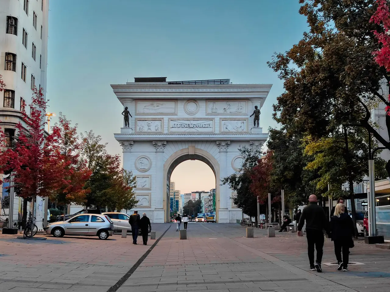The Porta Makedonija arch in Skopje at dusk in October 2022, where the poster of the JSP City Tour bus says the tour starts