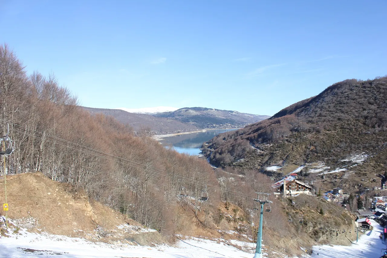 View down a chairlift line over bare woods to Lake Mavrovo, with resort hotels and patchy snow at the foot of the run