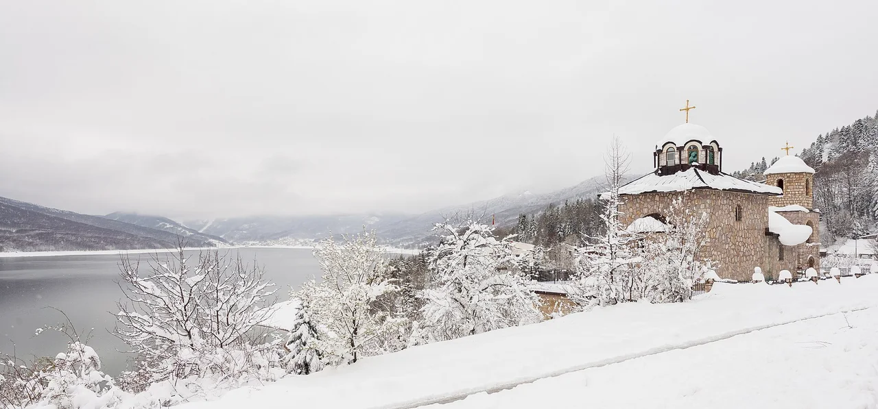 The stone church of Saints Cyril and Methodius in Mavrovi Anovi under heavy snow, with snow-laden trees and the grey lake below