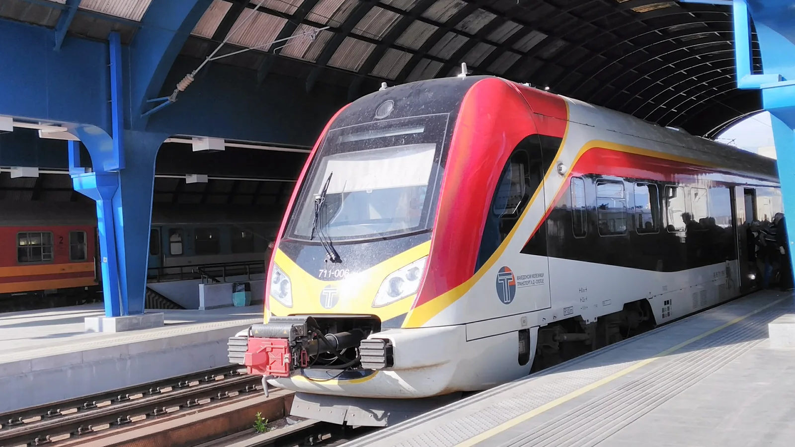 A red, white and yellow Macedonian Railways Transport railcar waiting under the blue arched platform roof of Skopje railway station