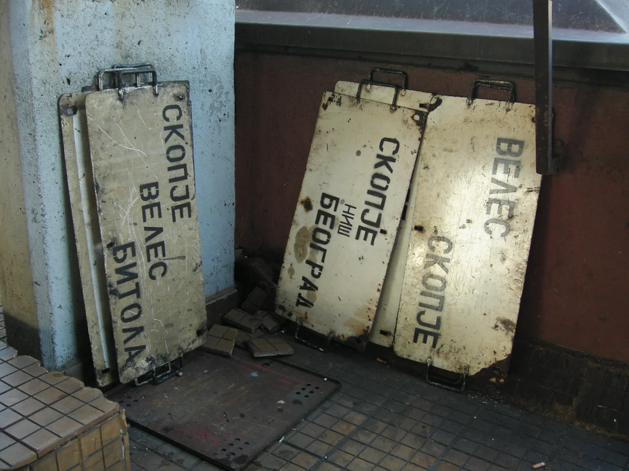 Three worn metal destination boards leaning in a corner of Skopje railway station, one reading Skopje - Veles - Bitola in Cyrillic