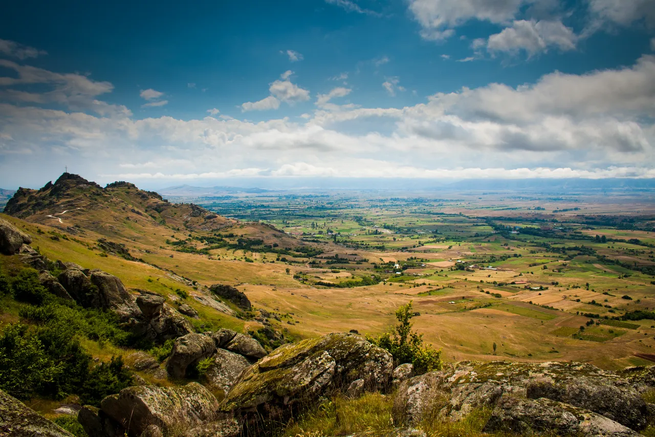 A wide view over the Pelagonia plain with patchwork fields, boulders in the foreground and a cross-topped hill on the left